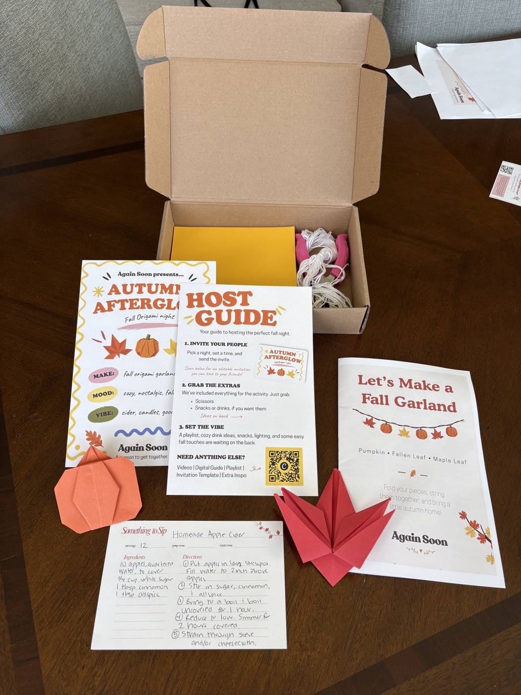 An open Again Soon box with origami paper and twine, beside the Autumn Afterglow theme card, host guide, garland instructions, a recipe card, and two finished origami pieces
